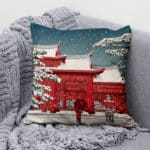 Coussin avec un bâtiment japonais rouge enneigé et un ciel bleu posé sur un canapé gris avec un plaid de la même couleur