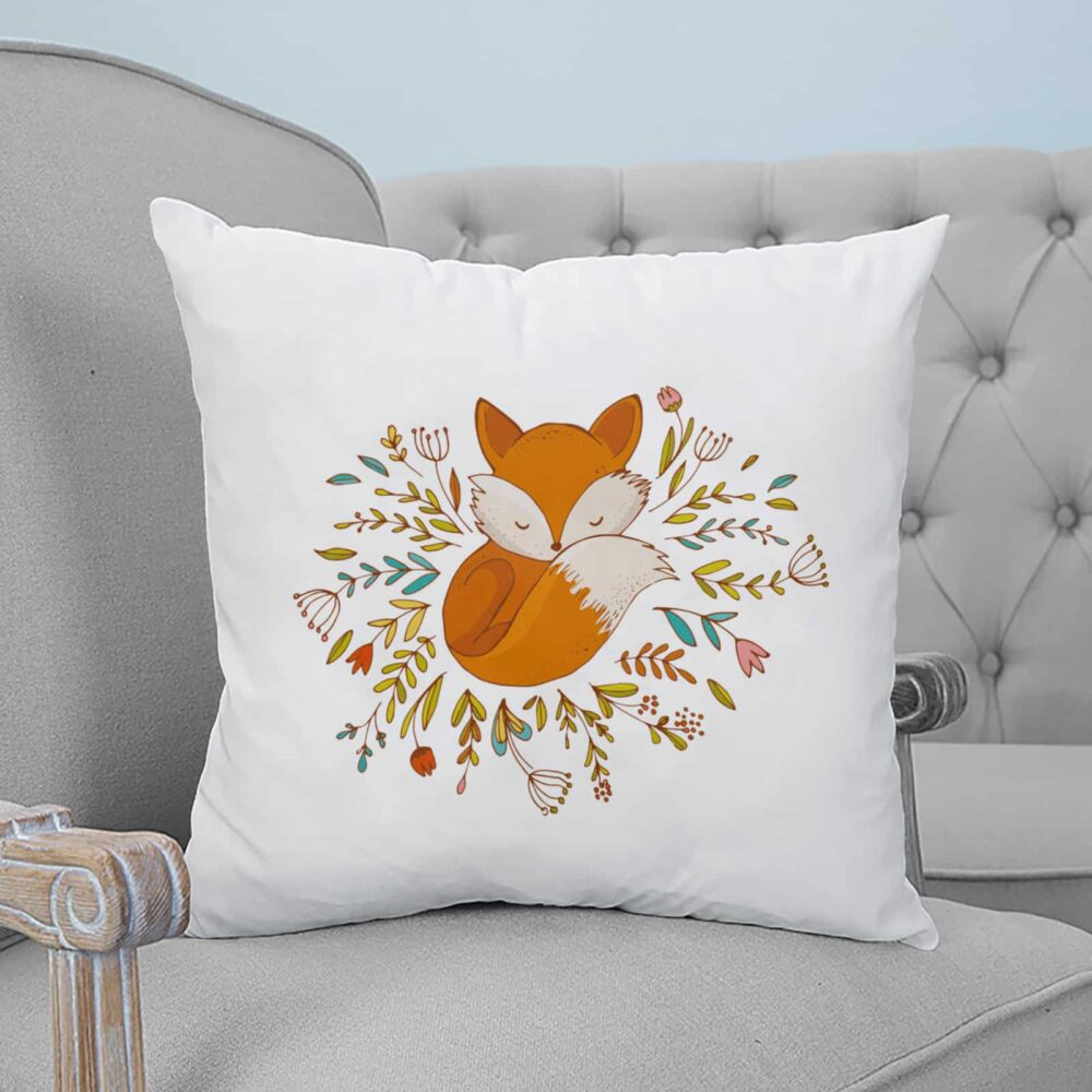 Coussin blanc carré avec un renard endormi et des fleurs dessinés dessus. Le coussin est posé sur un fauteuil gris