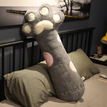 Gros coussin en forme de patte de chat grise avec des coussinets roses, posé sur un lit avec des draps et une bordure grise.