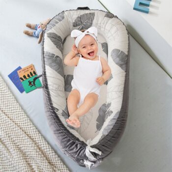 Coussin nid d'ange pour rassurer bébé. Il est gris avec un motif de feuilles. Il y a une petite fille habillée en blanc avec un bandeau qui rit à l'intérieur. Le coussin nid d'ange est posé sur le sol.