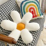 Coussin en forme de fleur blanc et jaune posé sur un canapé à carreaux noir et blanc avec un coussin au dessin d'arc-en-ciel posé dessus.