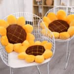 Coussin en forme de fleur de tournesols marrons au centre avec des pétales jaunes, posés sur des chaises en métal blanches sur un sol en parquet gris.