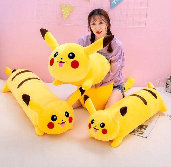 Femme à genou entourée de deux coussins Pikachu jaunes, longs et de formes cylindrique. Elle tient un coussin Pikachu dans ces bras identique aux deux autres