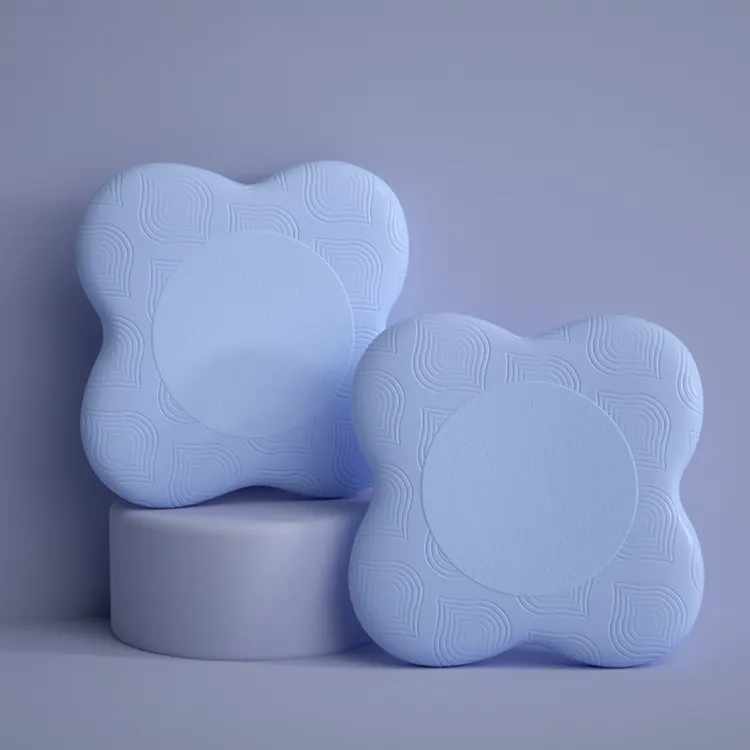 Deux coussins de yoga pour le genou et le coude de couleur bleu posés à la verticale sur un fond bleu, un est surélevé sur un objet de forme cylindrique également de couleur bleu.