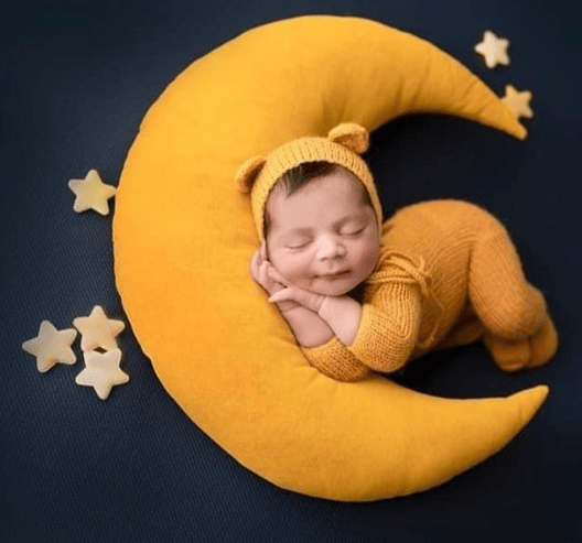 Joli coussin en forme de lune avec de petites étoiles en peluche jaunes moutarde, un bébé brun habillé en combinaison en maille moutarde dort au creux de la lune.