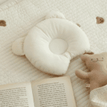 Joli coussin anti-tête plate en forme d'ourson blanc. Ile st posé sur un tapis d'éveil près d'un ourson et d'un roman.