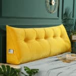 Coussin dossier de lit de lecture couleur moutarde. Est installé sur un lit devant un mur vert ou bleu canard. Coussin avec capitons.