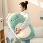 Femme debout tenant dans son bras gauche un coussin kawaii dinosaures vert clair de grande taille, le dinosaure a un cœur rose dessiné sur chaque joue, des dents blanches visibles, des mains dessinés sur le ventre, un ventre de couleur blanche, une crête jaune sur la tête, des petits pieds verts clairs visibles et une petite queue de reptile avec à l'extrémité une crête jaune.