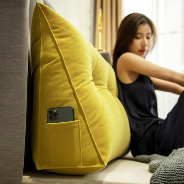 Femme dans une chambre assise sur un lit, son dos contre le coussin tête de lit jaune. Le coussin tête de lit jaune a une poche sur la gauche, à lintérieur de cette poche il y a un téléphone portable.