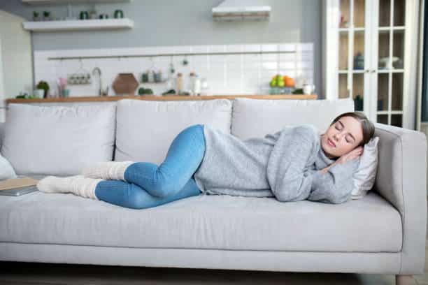 Femme apaisée en train de se reposer habillée avec un jean bleu et un pull-over gris couchée sur un canapé gris claire