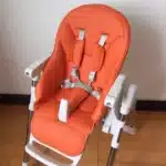 Coussin de chaise en cuir (Peg Perego Prima Pappa Zero3) de couleur orange avec un harnais 5 points attaché posé sur une chaise haute de couleur grise et blanche.