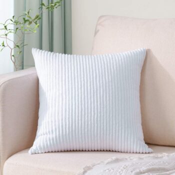 Coussin en velours blanc à rayures côtelées de forme carré, posé sur un canapé beige clair. On peut voir en arrière-plan une plante verte, un rideau vert pastel, un jeté de canapé blanc à motifs délicats est posé à côté du coussin.