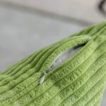 Coussin vert pomme en velours côtelé ayant une texture en relief (lignes parallèles) avec une fermeture éclair partiellement ouverte. La fermeture éclair est discrète, avec un curseur métallique argenté et un petit tire-zip blanc.