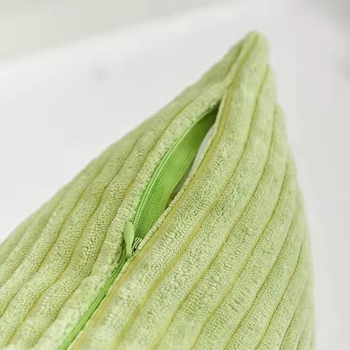 Coussin vert pomme en velours côtelé ayant une texture en relief (lignes parallèles) avec une fermeture éclair partiellement ouverte. La fermeture éclair est discrète, avec un curseur métallique argenté et un petit tire-zip blanc.