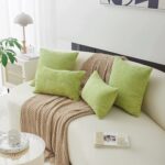 Quatre coussins en velours côtelé de couleur verte, deux de forme rectangulaire et deux carrés, apportant une touche naturelle et apaisante posés sur un canapé blanc, avec une texture douce et minimaliste. Autour il y a une table d’appoint ronde blanche avec une tasse et une soucoupe au design moucheté. Un livre avec une bougie décorative en forme de cubes blancs posé sur le canapé. Il y a aussi une lampe de table beige et blanche, au style moderne, placée sur un petit meuble avec une grille décorative. Une plante verte à feuilles larges ajoutant une touche de fraîcheur.