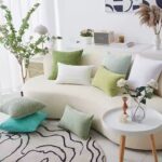 Salon moderne décoré avec plusieurs coussins en velours côtelé. La couleur des coussins est varié, il y a différentes nuances de vert (vert clair, vert olive, vert turquoise). Ils sont disposés sur le canapé, au sol et sur une table d’appoint. Il y a dans le salon une table d’appoint ronde blanche avec des pieds en bois, sur laquelle repose un vase en verre contenant des fleurs blanches et une bougie décorative en forme de cubes ; un tapis rond à motifs abstraits noirs et blancs, ajoutant une touche artistique ; un meuble d’appoint avec une grille décorative, surmonté d’une lampe de table blanche et de petits objets déco ; une étagère murale en arrière-plan, avec un haut-parleur Marshall, des livres et une bougie ; un grand vase blanc avec des pampas beiges, apportant une touche naturelle et bohème. une grande plante verte en pot près de la fenêtre, ajoutant de la fraîcheur.