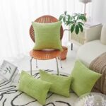 Ensemble de quatre coussins verts en velours côtelé avec une texture en relief (lignes horizontales) disposés dans un intérieur moderne et lumineux. Un coussin carré posé sur une chaise design en bois avec dossier ajouré, au design élégant, trois autres coussins rectangulaires sont disposés sur un tapis et un canapé. A côté on peut apercevoir un canapé clair et arrondi, avec une couverture en maille beige, une table d’appoint blanche avec une tasse et une assiette, une plante verte en pot, un petit meuble blanc avec un détail en rotin, un porte-revues métallique avec un magazine sur la nature, on peut voir un tapis à motif abstrait noir et blanc.