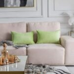 Deux coussins rectangulaires verts pomme en velours côtelé avec des lignes horizontales, placés sur un canapé beige clair avec un design moderne et minimaliste. Une revue ou un magazine avec une couverture en noir et blanc repose sur une couverture grise en maille tricotée. Une table d'appoint dorée avec un plateau en verre est placée à côté du canapé. Sur la table, une boîte cadeau emballée et une décoration en plumes blanches ajoutent une touche sophistiquée.