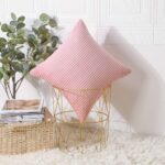 Coussin en velours rose à rayures côtelées de forme carré et légèrement bombée, posé en diagonale et de manière élégante sur une table d’appoint dorée avec une structure géométrique ajourée. A côté de la table il y a un panier en osier naturel, rempli de magazines, on peut voir en arrière-plan, une plante verte avec de grandes feuilles. La table, la plante verte et le panier sont posés sur un tapis en fausse fourrure blanche.