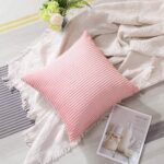 Coussin carré en velours côtelé de couleur rose, de forme carrée avec des lignes verticales régulières. Le coussin est légèrement bombé, bien rempli. Le coussin est posé sur un plaid blanc cassé à motifs géométriques discrets avec des franges sur les bords. On aperçoit une revue ou une affiche en bas à droite, illustrant des pots de lait avec le titre "MILK IS SWEET", renforçant l’ambiance cocooning. À côté du coussin, il y a un petit bouquet de fleurs artificielles avec des roses blanches et des feuillages verts. Tout est posé sur un tapis rayé dans des tons gris et beige.