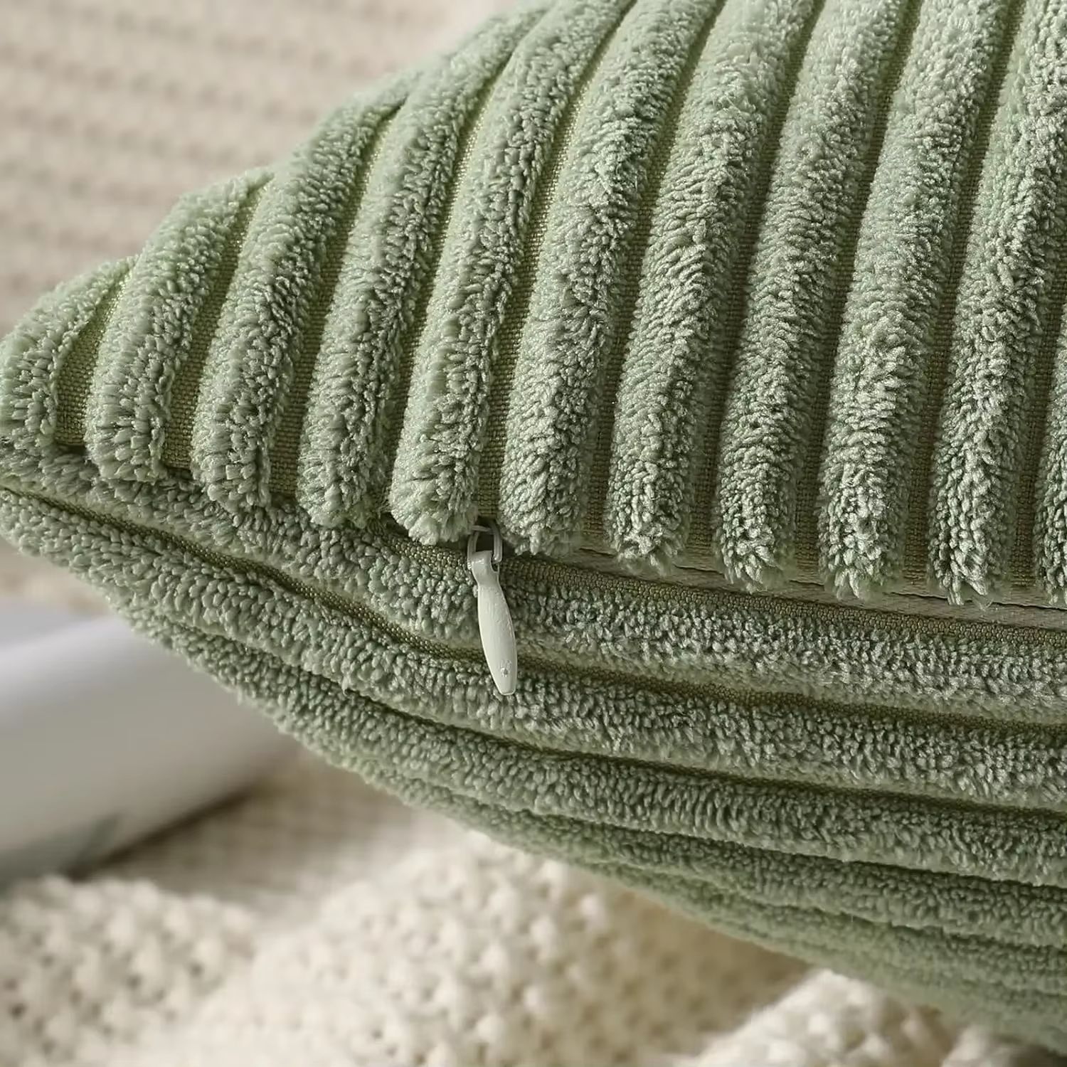 gros plan d’un coussin en velours côtelé vert sauge avec des rayures en relief bien marquées, mettant en avant la texture du tissu et la fermeture éclair dissimulée. La fermeture éclair est placée discrètement sur la couture latérale du coussin et assortie à la couleur du tissu, ce qui la rend presque invisible. L’arrière-plan flou laisse apparaître un plaid tricoté beige clair.