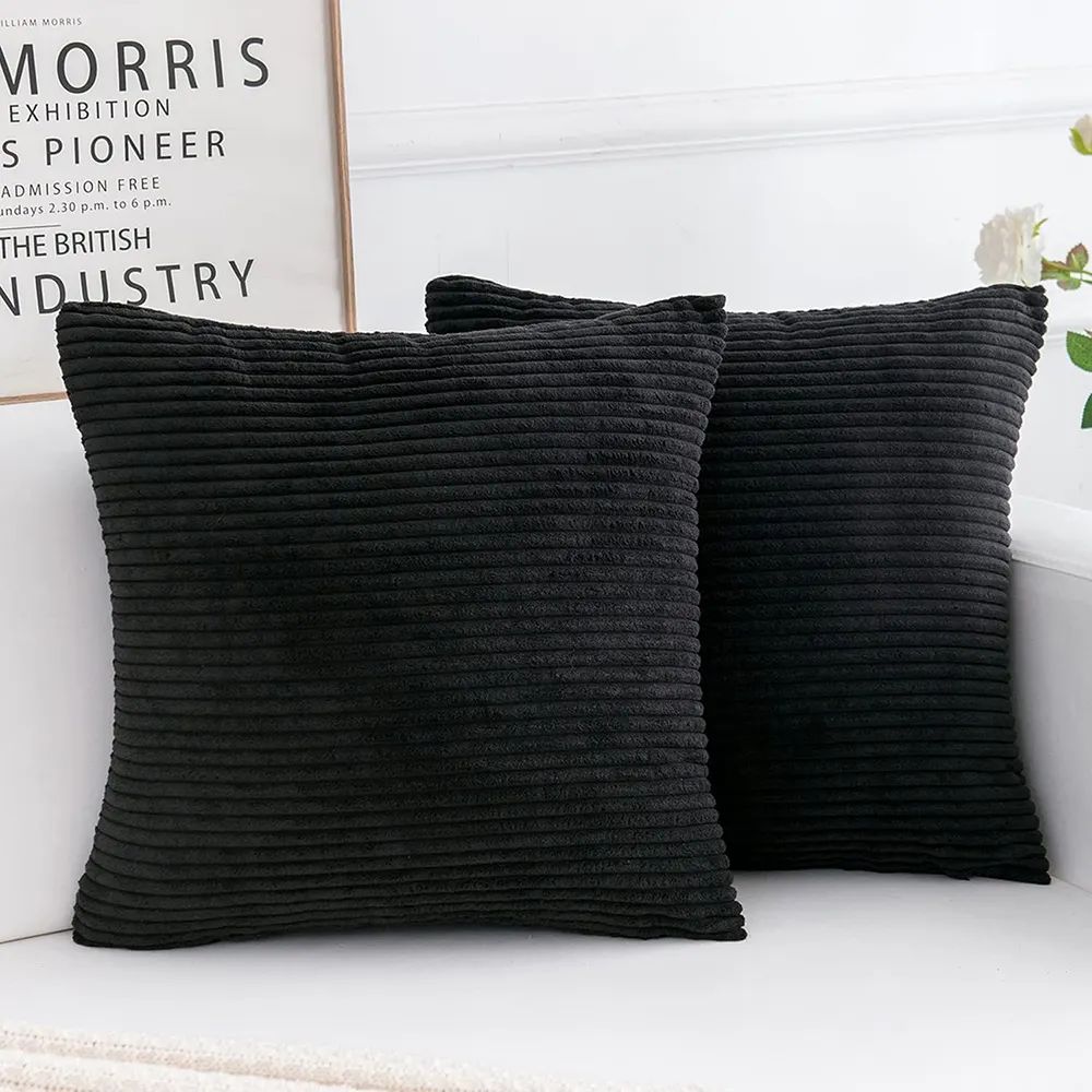 Deux coussins en velours côtelé noir de forme carrée disposés sur un canapé blanc dans un intérieur élégant et épuré. Un plaid en tricot beige est posé sur l’accoudoir. Un cadre mural avec une inscription sur le design textile et William Morris. Quelques fleurs blanches en arrière-plan, apportant une douceur visuelle.